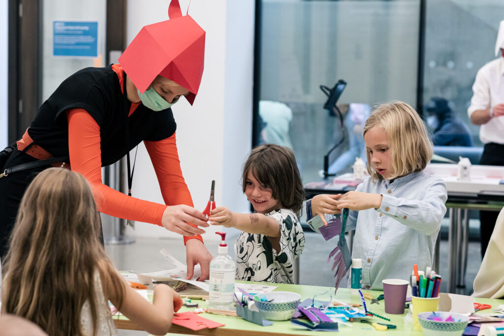 Photo of some children standing busily around a table covered with craft materials. One child is smiling whilst being handed a pair of scissors by a woman wearing a red paper hat in the shape of a horse head.