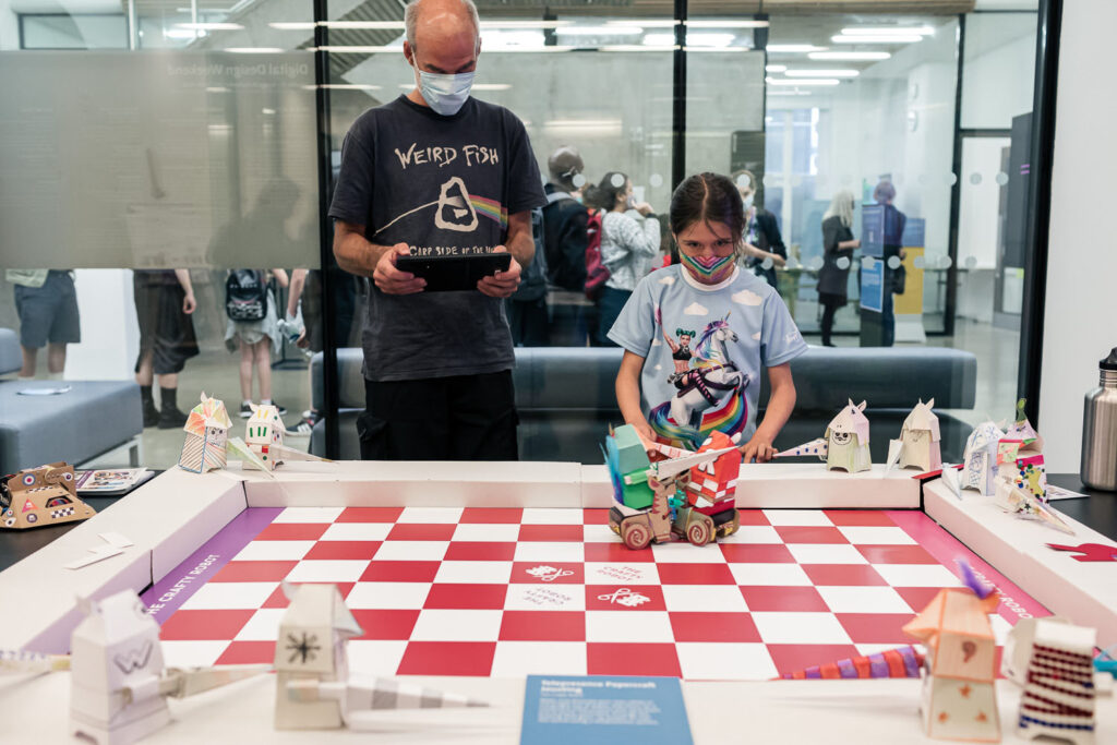 Photo of a man in his 50s and a girl of about 10 standing behind a large table holding iPads. On the table is a red and white chessboard pattern on which two robot unicorns carrying paper knights are colliding. Around the edge are other hand decorated paper knights.