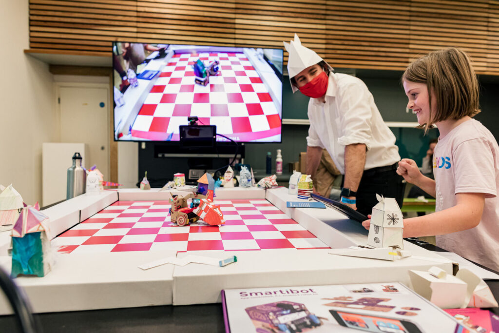 Photo of the table with the chessboard playing surface and robot unicorns with paper knights. There is a smiling child and a man wearing a white paper hat in the shape of a horse head standing to one side and you can see at the end of the table a large screen shows what is happening on the playing surface.