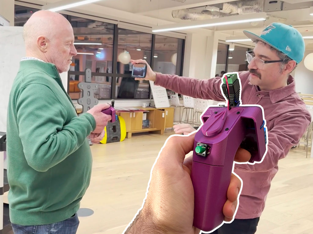 Photo of an older man holding a gun shaped device pointing it at a younger man who is handing the older man a phone. Composited on top is an image of the gun shaped device showing it to have a tactile pointer on the top.